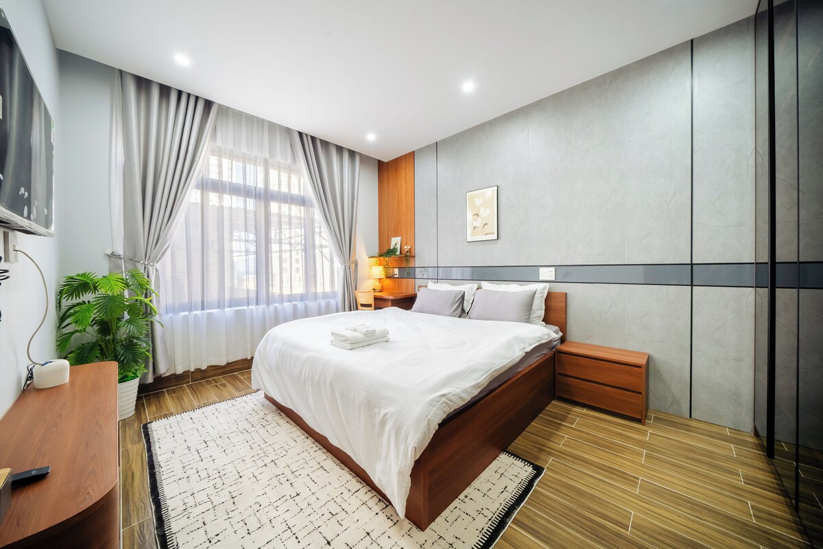 A modern hotel room with a large bed, a bedside table, a nightstand, a potted plant, and a large window with curtains.
