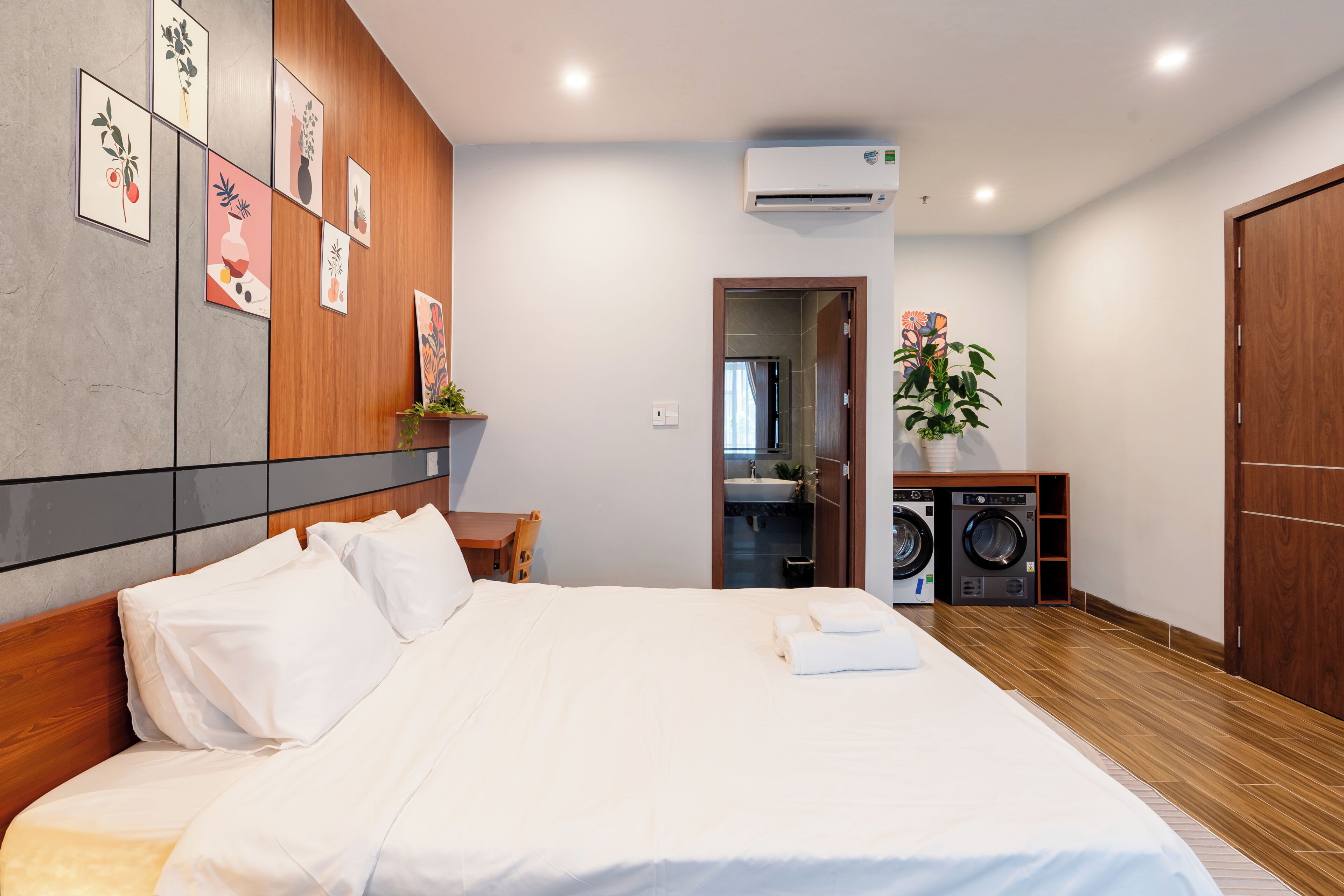 Habitación de hotel con cama, cabecera de madera, aire acondicionado empotrado, lavadora y baño visible a través de una puerta abierta.