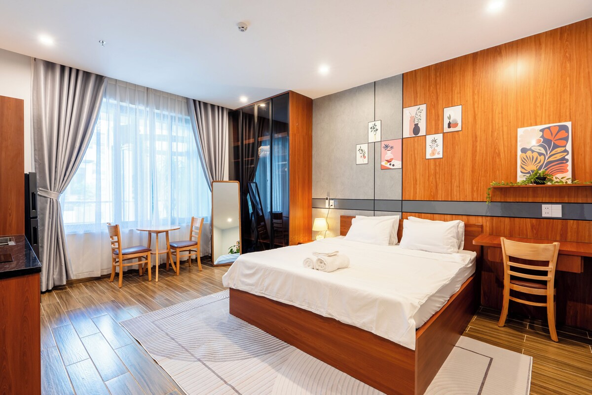 A modern hotel room with a large bed, a desk, a chair, and a dining area.