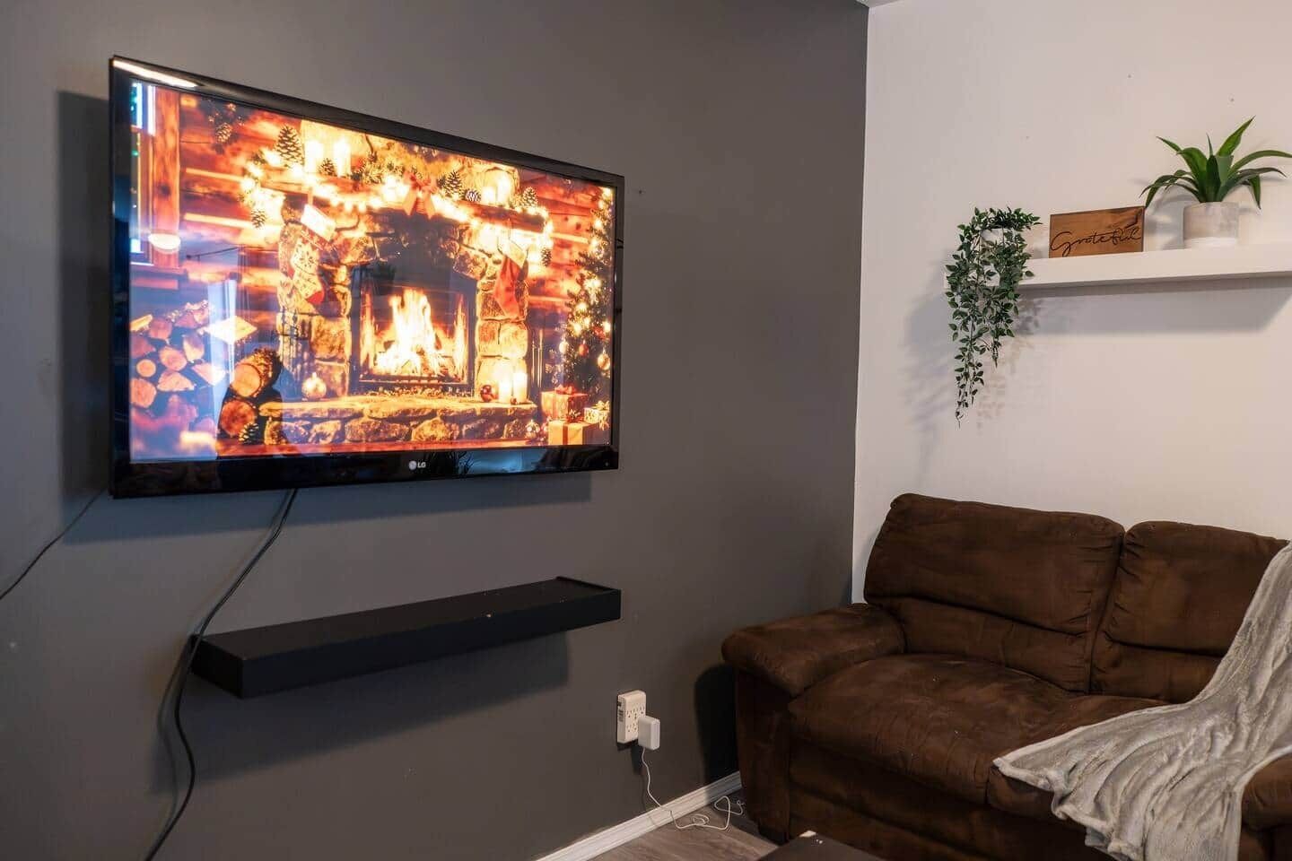 Una TV a schermo piatto montata a parete, un divano marrone, una piccola mensola con una pianta e una presa elettrica.