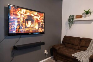 Una TV a schermo piatto montata a parete, un divano marrone, una piccola mensola con una pianta e una presa elettrica.
