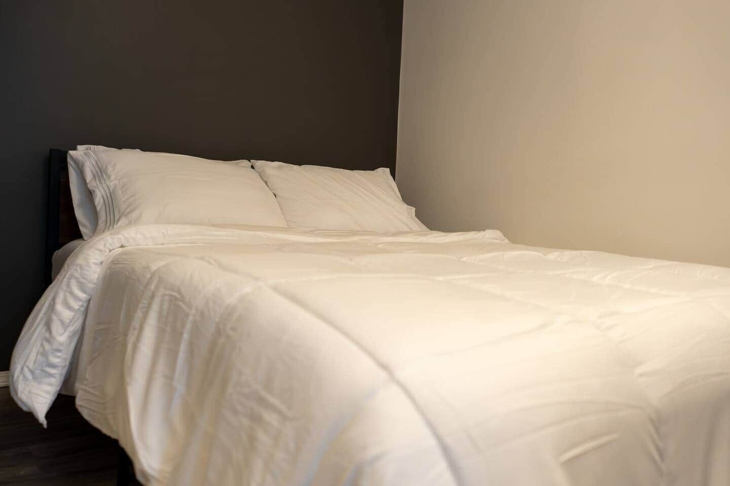 A neatly made bed with white bedding in a room with a dark wall and a light-colored floor.