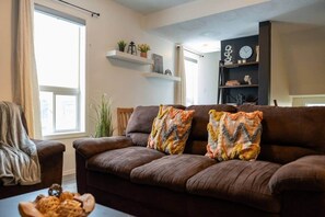 Un soggiorno con un divano marrone, due cuscini decorativi a motivi, un tavolino con una ciotola di pasta e una libreria con oggetti ornamentali.