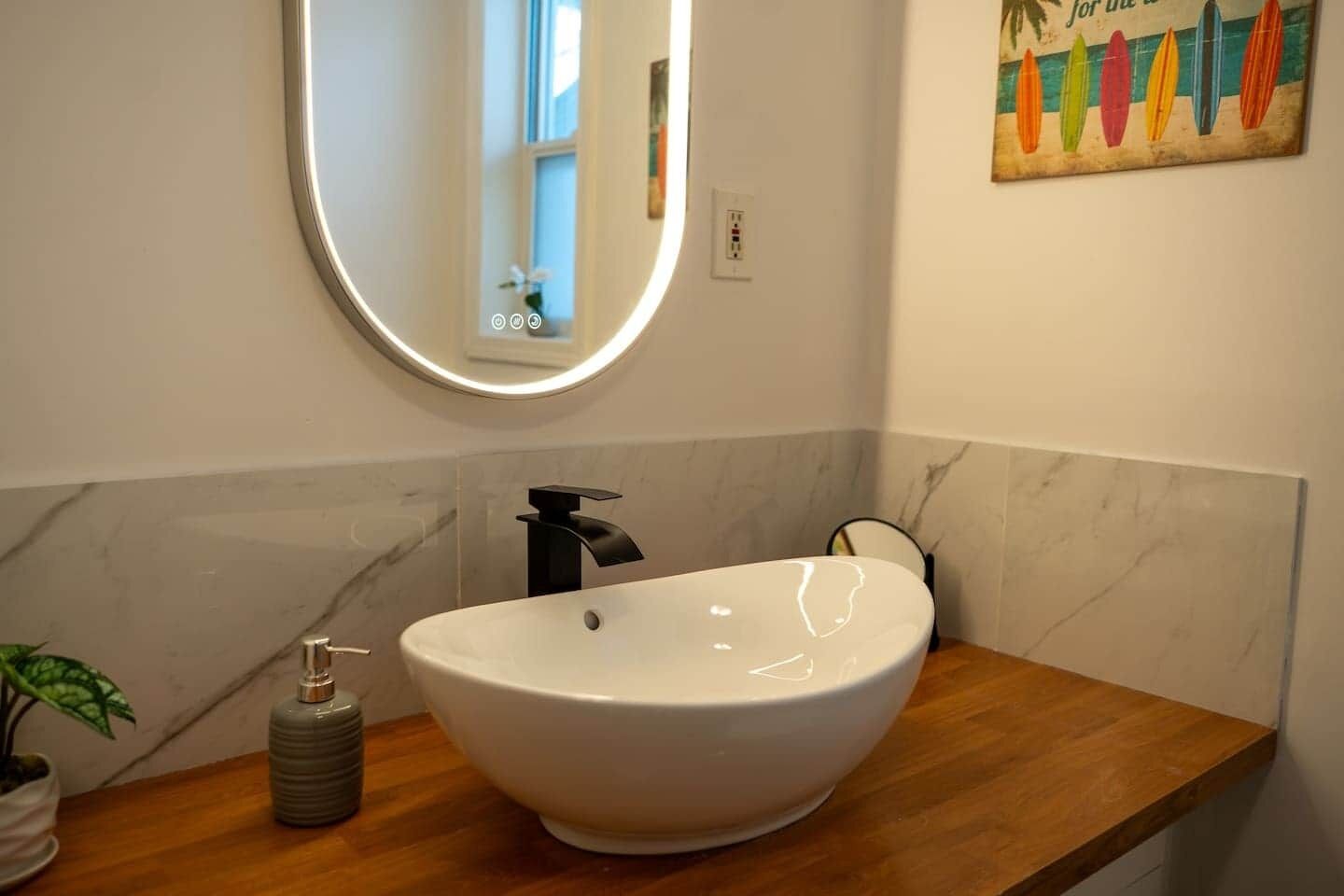 Ein Badezimmer mit einem weißen Waschbecken, einem runden Spiegel, einer Holztischplatte und einer kleinen Topfpflanze.