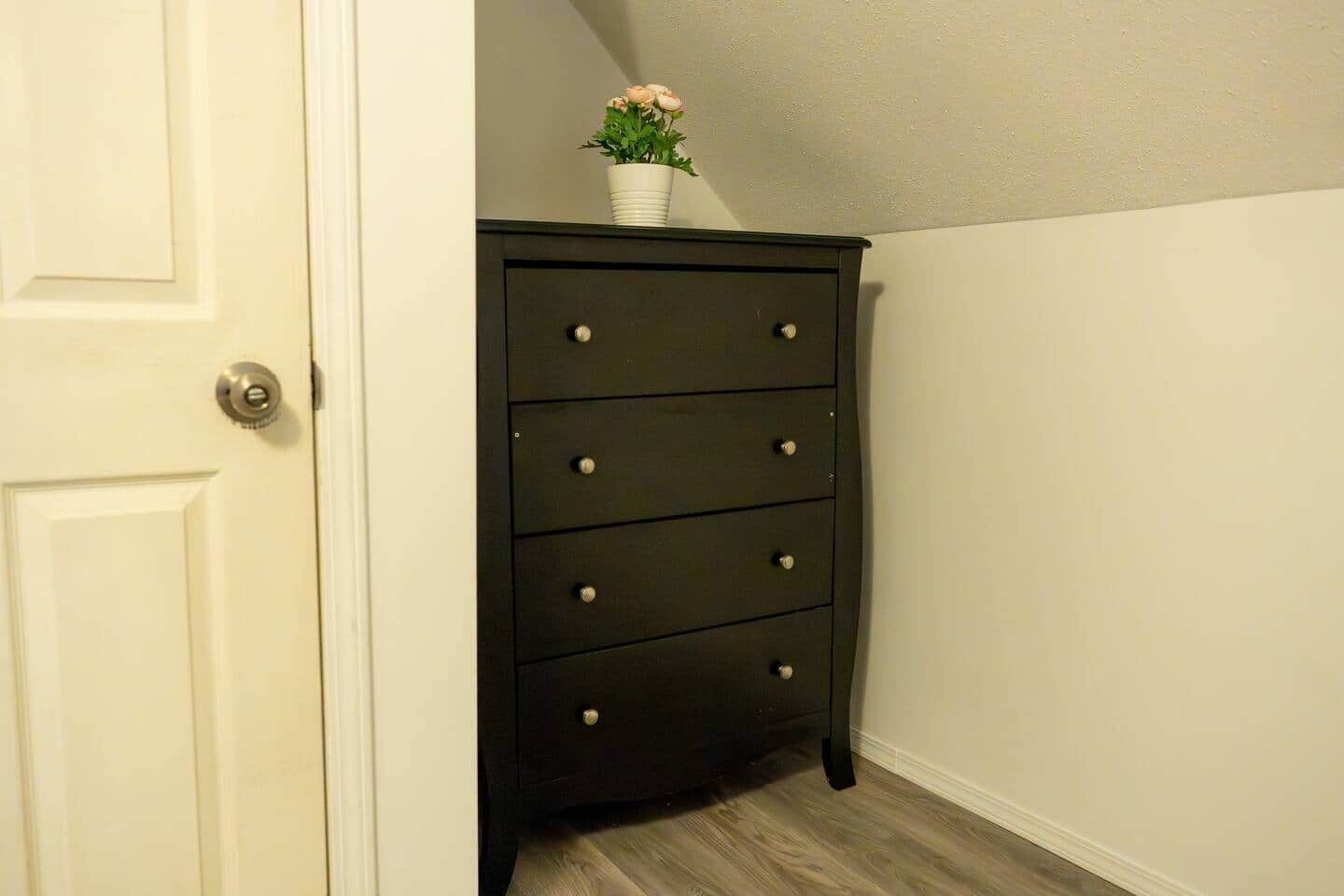 A dark wooden dresser with four drawers and a small potted plant on top, positioned against a white wall with a door to the left.