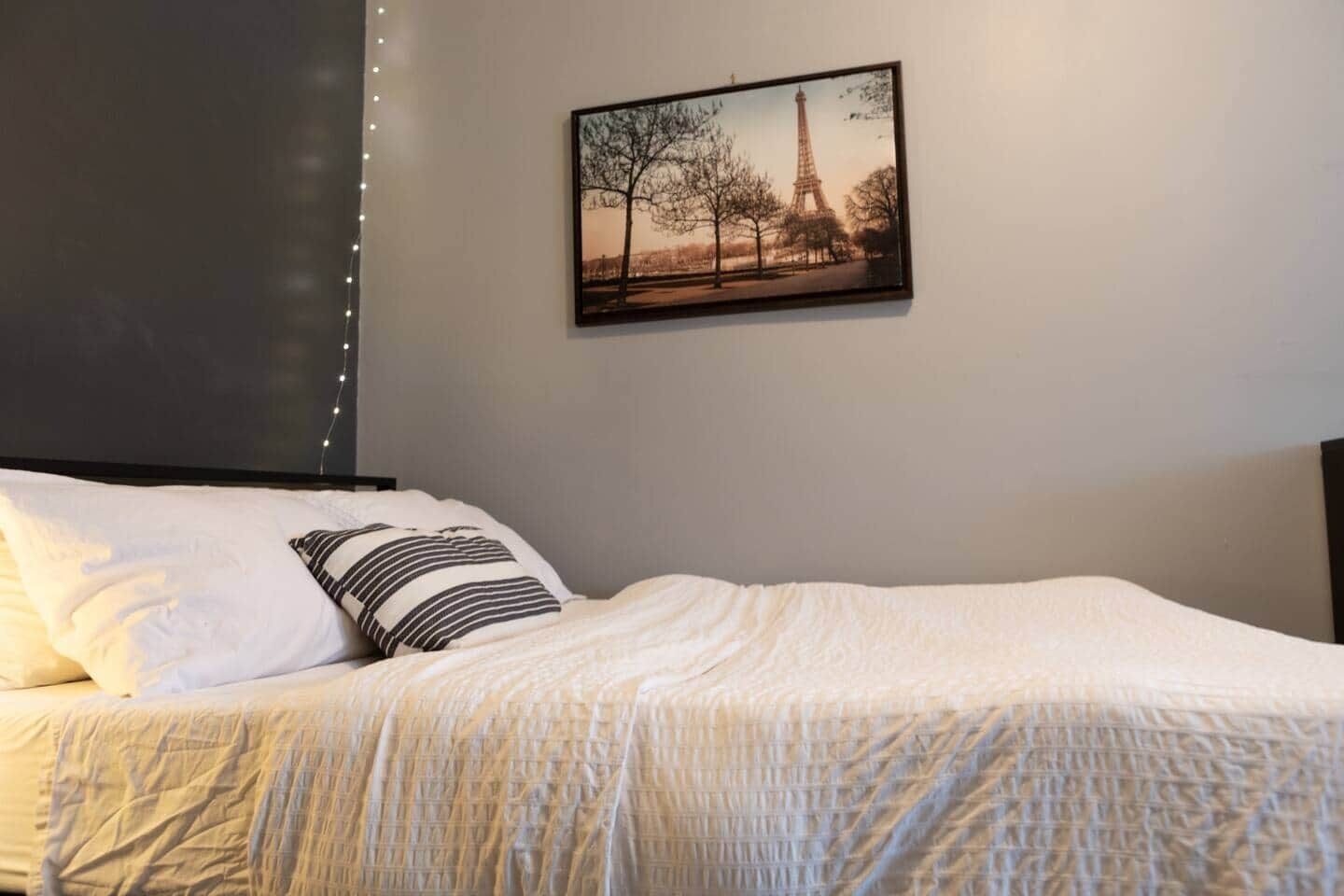 Una cama bien hecha con ropa blanca y almohadas a rayas, un cuadro con la Torre Eiffel en la pared y luces decorativas a la izquierda.