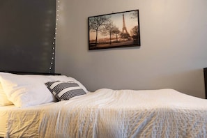 Ein ordentlich bezogenes Bett mit weißer Bettwäsche und gestreiften Kissen, ein gerahmtes Bild vom Eiffelturm an der Wand und Lichterketten auf der linken Seite.