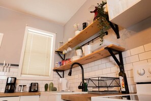 Eine Küche mit weißen Fliesen, einem Fenster mit Jalousien, Holzregalen mit Küchenutensilien, einem schwarzen Wasserhahn und einer Weinflasche.