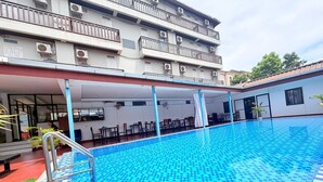 A swimming pool with a checkered pattern, surrounded by a covered seating area with tables and chairs, and a multi-story building with balconies in the background. - Cozy Corner Hotel (Vang Vieng)