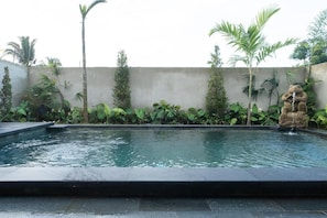 Ein rechteckiger Pool mit Steinbordierung, umgeben von tropischen Pflanzen und einer Betonmauer.