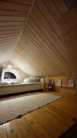Une chambre à coucher aménagée dans un espace sous les toits, avec un plafond en pente, un lit recouvert d’une couvre-lit à motifs, une petite table de chevet, une commode et une fenêtre offrant une vue panoramique.