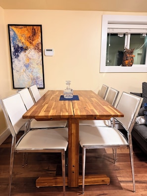 A dining table with a wooden top and metal legs, surrounded by white chairs, in a room with a large abstract painting on the wall and a window with an owl decoration.