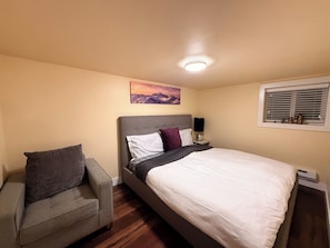 A bedroom with a bed, a nightstand, a lamp, a chair, a painting, and a window with blinds.