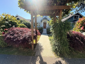 A house with a wooden pergola entrance, a pathway leading to the front door, and a well-maintained garden with various plants.