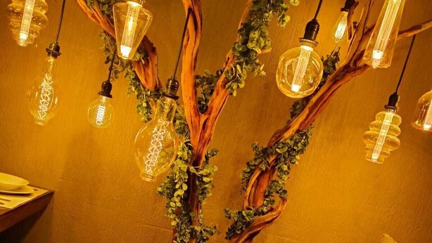 Estructura en forma de árbol con bombillas colgantes y follaje, ubicada en una esquina de una habitación con piso de madera y un banco acolchado.