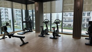 A gym with various exercise equipment, large windows, and a view of residential buildings. - Aravali Hill CherisingBeauty 1BHK CP3Flower Valley (Gurugram)