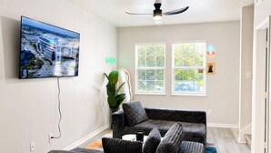 A living room with a flat-screen TV mounted on the wall, a ceiling fan, a large window with blinds, and a plant. - Space City 4BR - 7 Min to NRG Stadium (Houston)