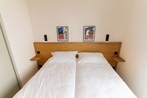 Een tweepersoonsbed met witte lakens, twee wandspeakers en twee ingelijste posters op het hoofdeinde.