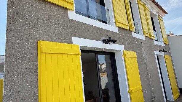 Un edificio con contraventanas amarillas, un balcón y una puerta que da a un espacio interior.