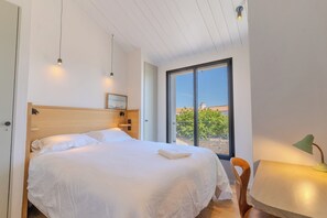 A bedroom with a large bed, a wooden headboard, a bedside table with a lamp, and a window with a view of buildings and trees.