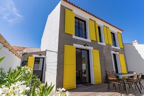 Une maison à deux étages avec un balcon, un coin repas comprenant des chaises et une table, et un jardin agrémenté de fleurs blanches.