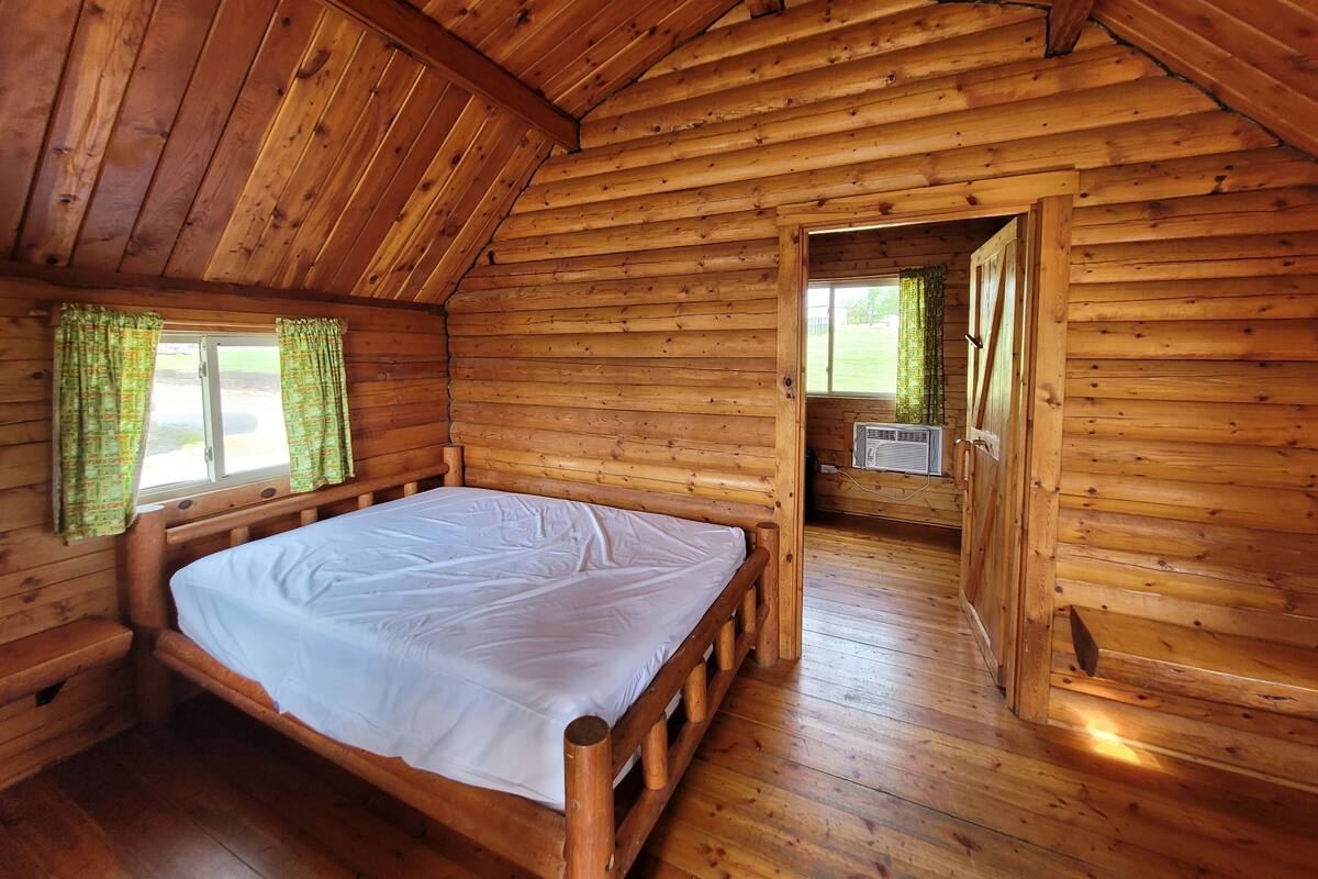A wooden cabin with a bed, a window with curtains, and a door leading to another room.