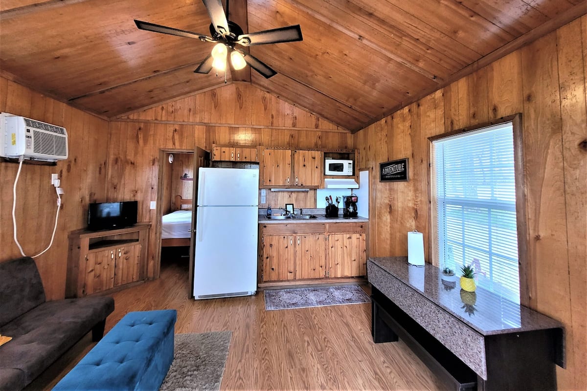 A cabin interior with a kitchenette, a ceiling fan, a television, a sofa, and a window with blinds.