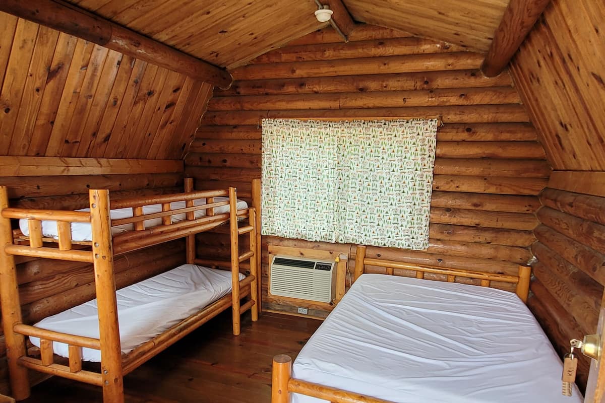 A cabin with wooden walls, two bunk beds, a window with patterned curtains, and a small air conditioning unit.