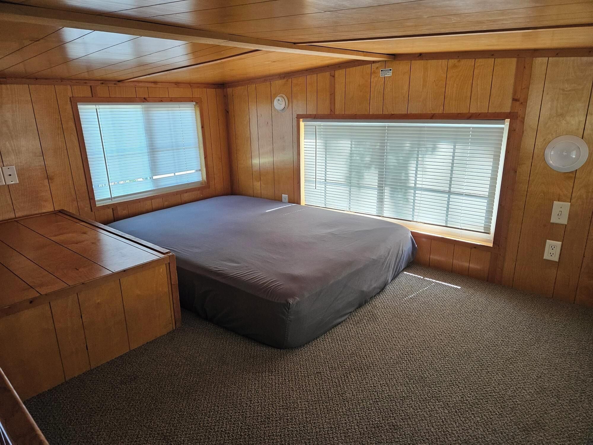 Een compacte kamer met een bed, een klein bureau en twee ramen met jaloezieën.