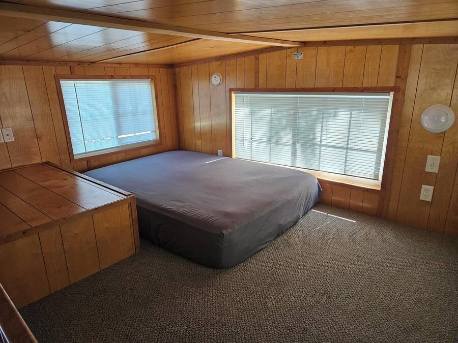 Een compacte kamer met een bed, een klein bureau en twee ramen met jaloezieën.