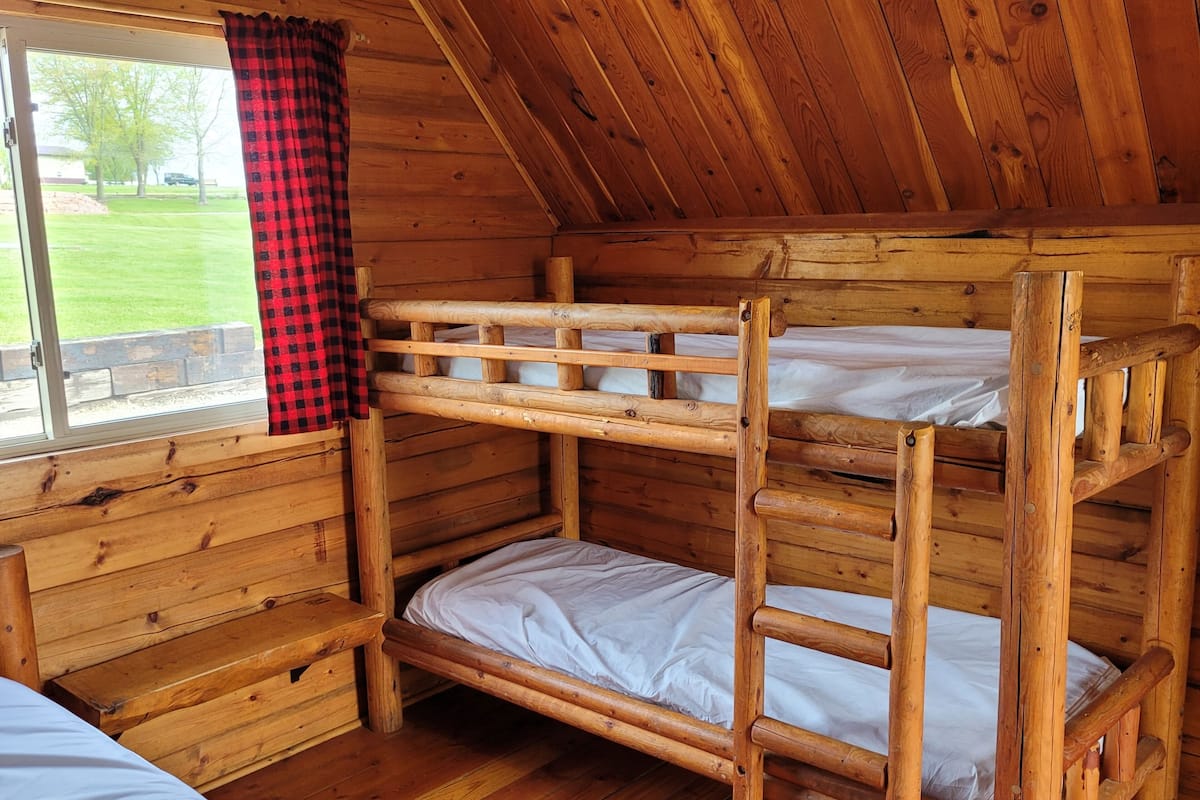 A bunk bed room with wooden walls and a window with a red and black checkered curtain.