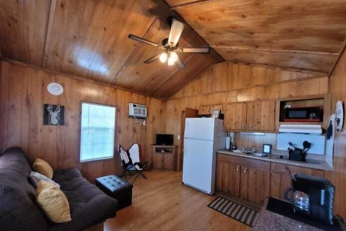 A cabin interior with a wooden ceiling, a ceiling fan, a kitchenette, a sofa, and a window.