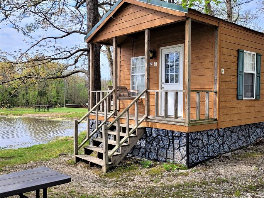 A wooden cabin with a porch and stairs, situated near a body of water and trees.