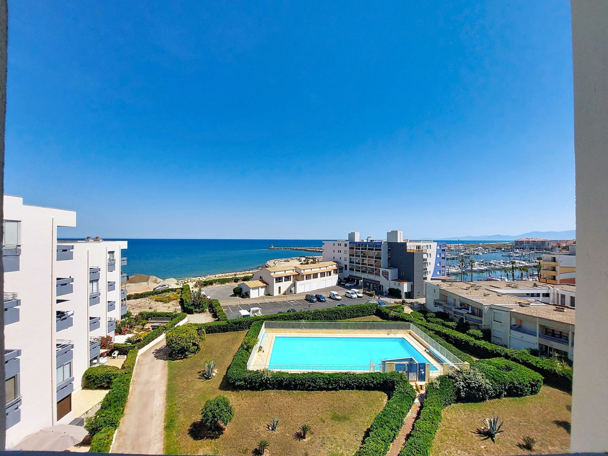 A coastal hotel with a swimming pool, multiple buildings, and a marina in the background.