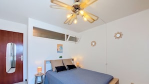 Ein Schlafzimmer mit einem Bett, einem Deckenventilator, einem kleinen Tisch mit einer Lampe, einem Spiegel und einer Wandleuchte.