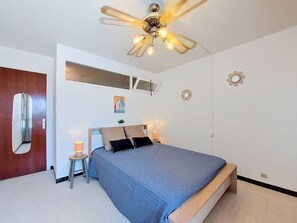 A bedroom with a bed, a ceiling fan, a small table with a lamp, a mirror, and a wall-mounted light fixture. - Port Saint Ange by Interhome (Le Barcarès)