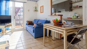 Una sala de estar con sofá, televisor, mesa de comedor con sillas, zona de cocina con armarios y balcón con vistas.