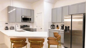 Una cocina moderna con armarios grises, electrodomésticos de acero inoxidable, una encimera blanca y cuatro taburetes marrones.