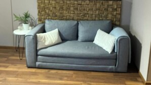Ein graues Sofa mit einem gewobenen Kopfteil, ein kleiner weißer Nachttisch mit einer Pflanze und ein Holzboden.