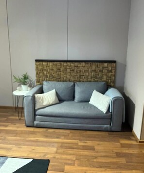 A grey sofa with a woven headboard, a small white table with a plant, and a wooden floor.