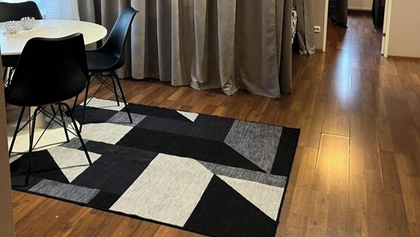 Ein modernes Zimmer mit Esstisch, Stühlen, einem gemusterten Teppich und einem großen Fenster mit Vorhängen.