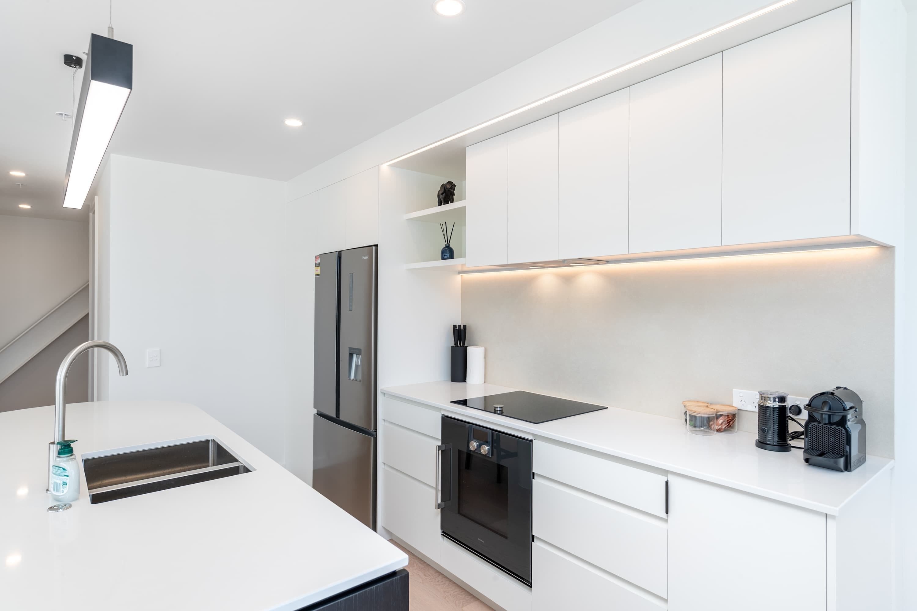 A modern kitchen with white cabinetry, a built-in oven, a stainless steel refrigerator, and a countertop with a coffee machine and other appliances.