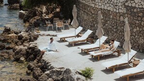 Ein Liegebereich am Strand mit Liegestühlen und Sonnenschirmen, einer Steinmauer und einer felsigen Küstenlinie.