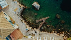 Luftaufnahme eines Küstenhotels mit klarem Wasser, einer felsigen Küstenlinie und einer Holzterrasse.