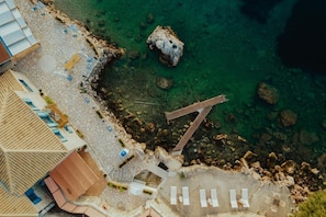 An aerial view of a coastal hotel with clear waters, a rocky shoreline, and a wooden deck. - Deluxe Studio - Sea Mansion Lefkada (Lefkada)