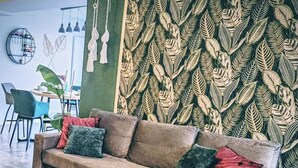 Uma sala de estar com um sofá, um banquinho redondo, um tapete vermelho e uma parede estampada com folhas.