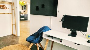 Quarto com escrivaninha, cadeira, televisão e cozinha ao fundo.