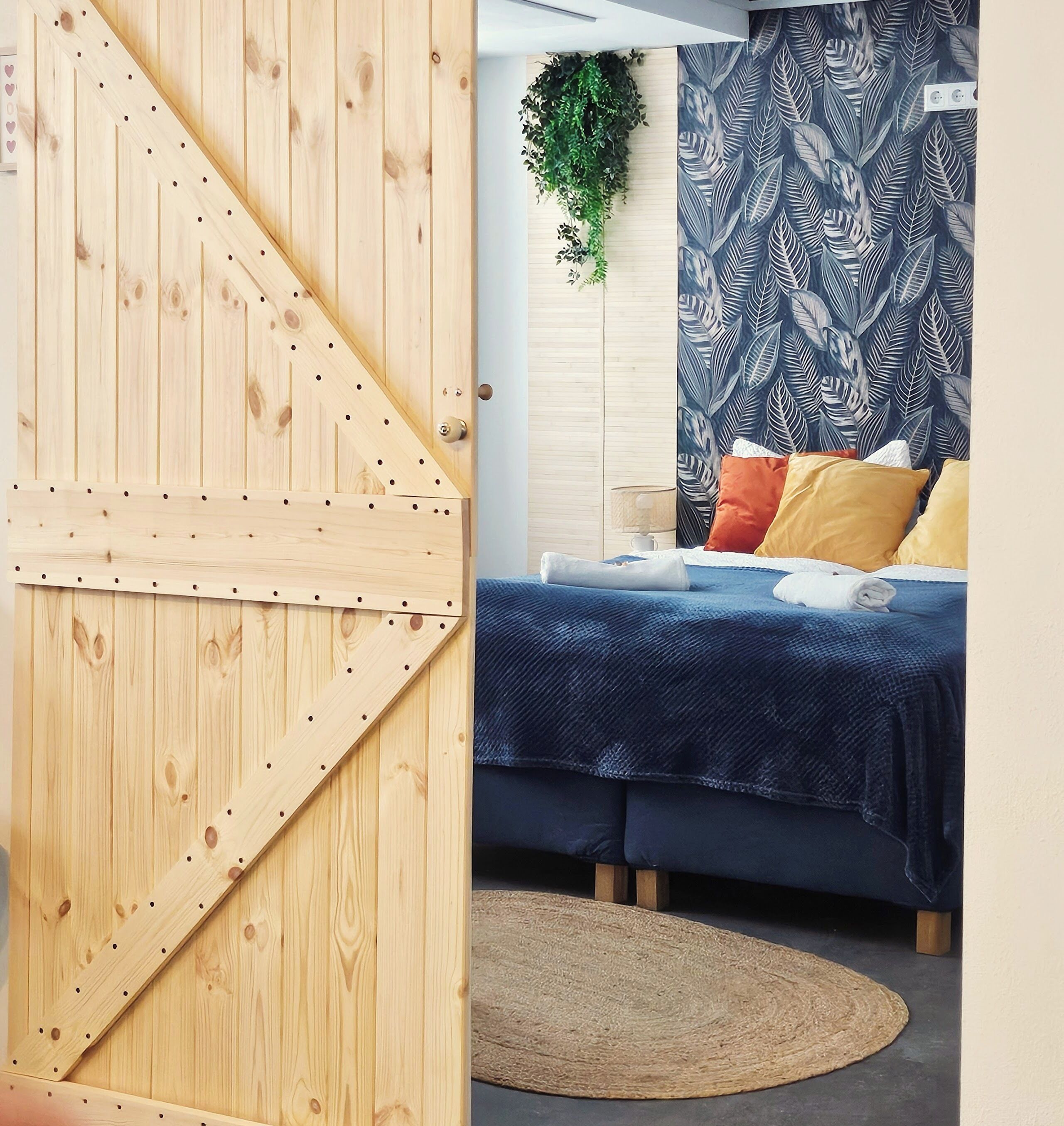 A room with a wooden door, a bed with a blue bedspread, a patterned wall, and a woven rug.