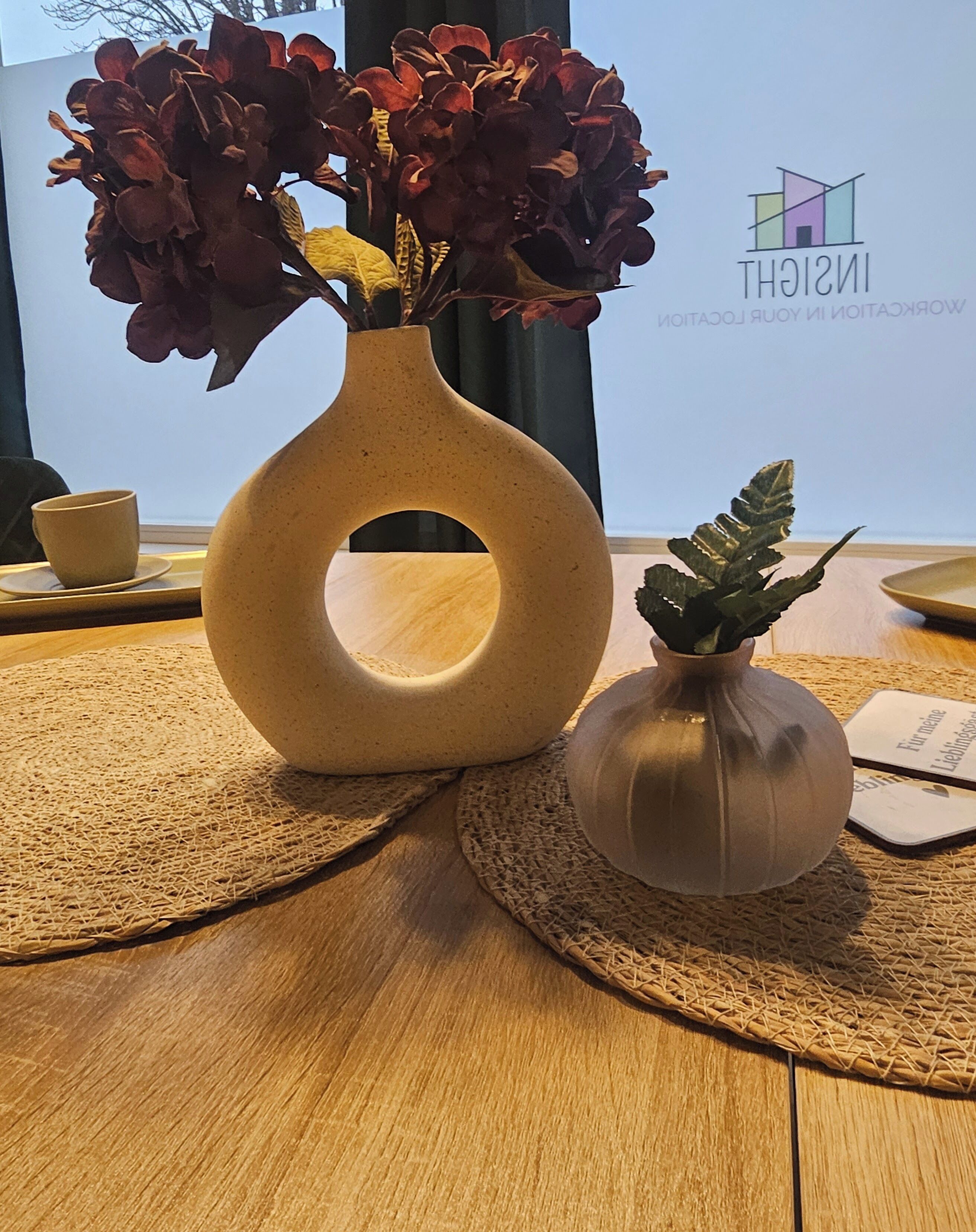 Ein runder Tisch mit einer gewebten Tischmatte, einer Vase mit getrockneten Blumen und einer kleineren Vase mit einer Grünpflanze.