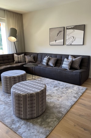 Ein modernes Wohnzimmer mit einer Eckcouch, Poufs, einem Teppich und zwei gerahmten Bildern an der Wand.